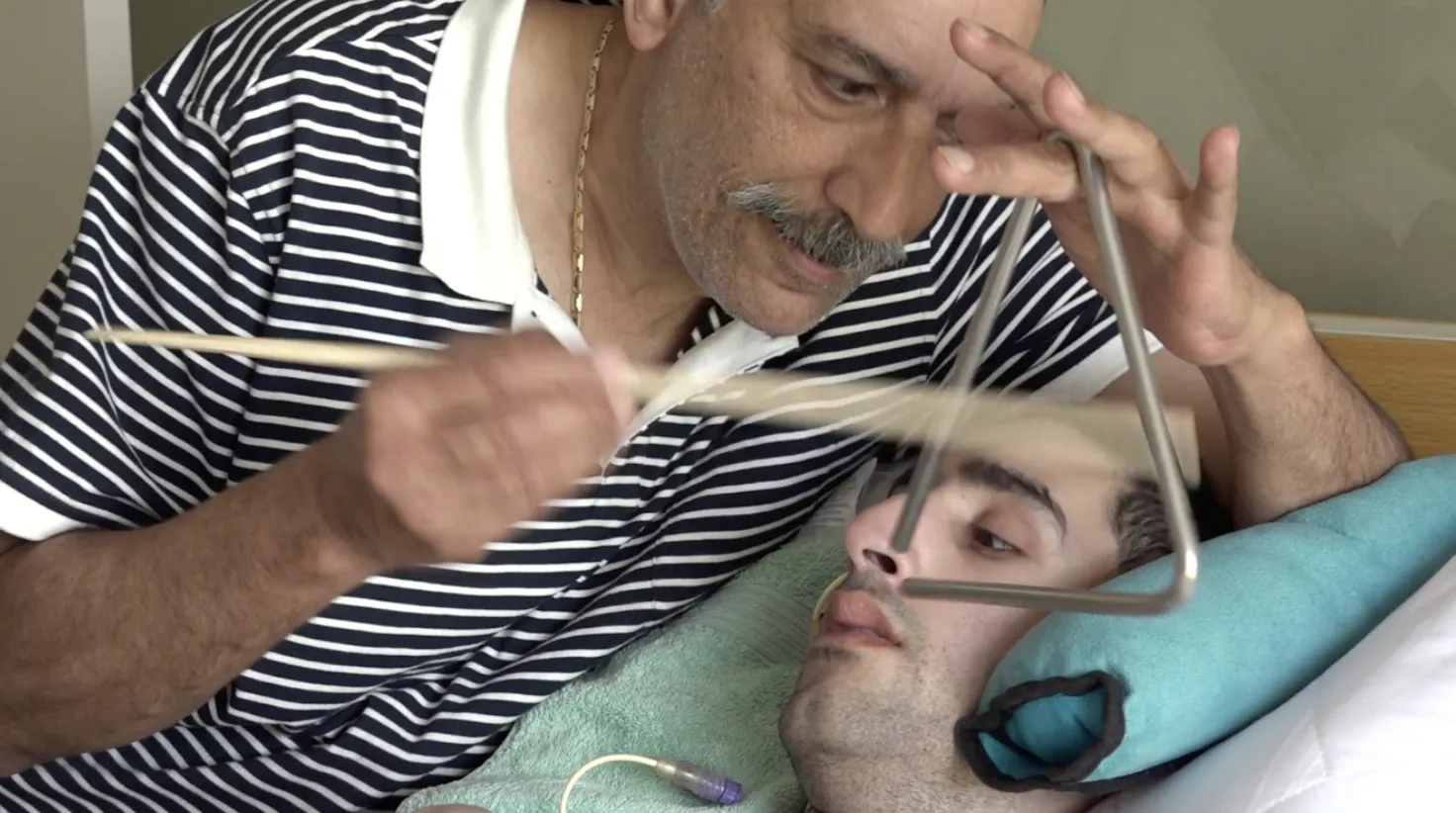 A carer plays a triangle close to a young man lying in bed, who watches intently
